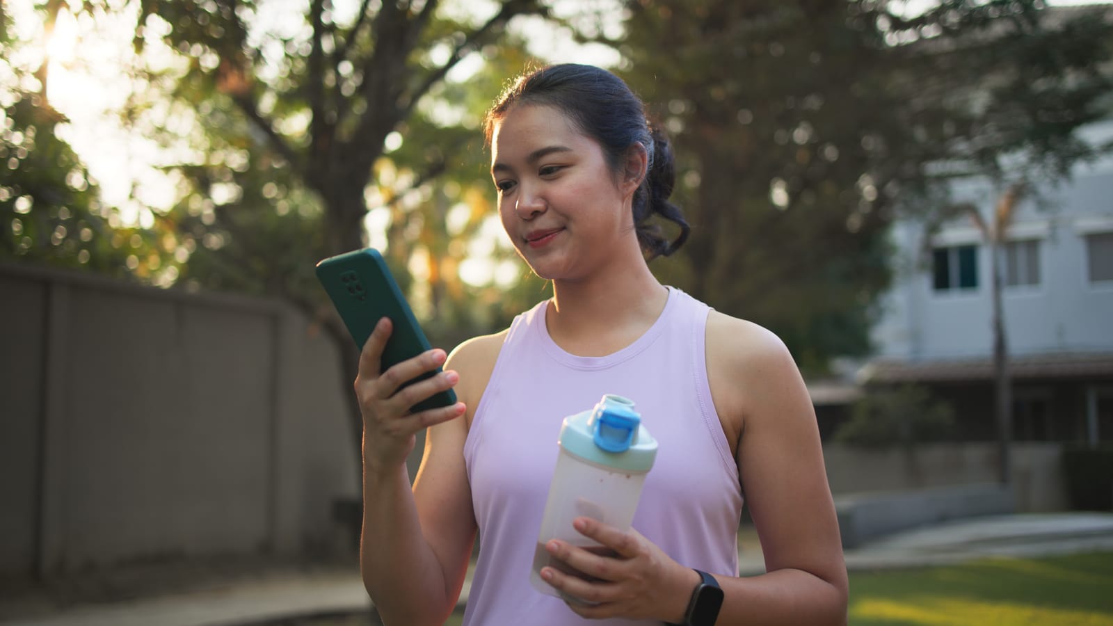 A woman checking her phone outdoors, holding a shaker bottle after a workout